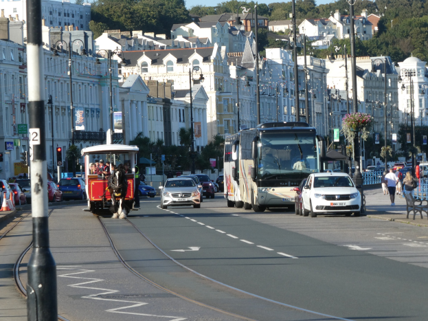 Eine belebte Stadtstraße mit hohen Gebäuden, Fahrzeugen, Fußgängern, Gehwegen, Laternen, Verkehrsampeln, Schildern, Bäumen und einem klaren blauen Himmel.