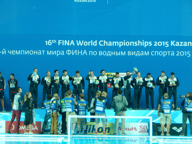 Gruppe von Menschen auf einem Schwimmbeckenrand mit Kameras, einem Netz im Vordergrund und einem "FINA World Championships 2015 Kazan"-Banner im Hintergrund.