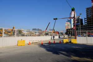 Eine Baustelle mit zahlreichen Verkehrskegeln, eine Person auf der Straße umgeben von Absperrungen, ein Zaun, ein Schild, ein Straßenpfahl, ein Verkehrssignal, zusätzliche Pfähle, Gebäude mit Fenstern, Kräne, Bäume und ein bewölkter Himmel.