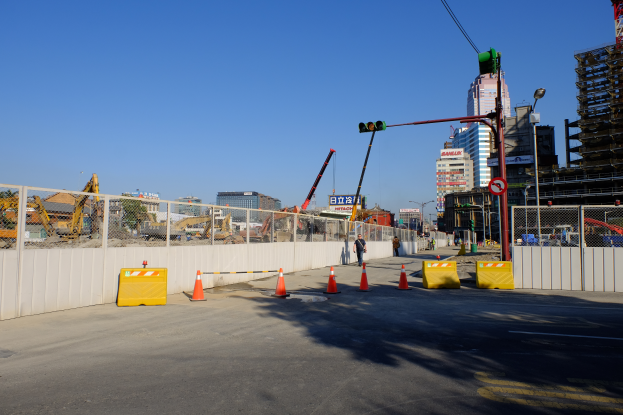Eine Baustelle mit zahlreichen Verkehrskegeln, eine Person auf der Straße umgeben von Absperrungen, ein Zaun, ein Schild, ein Straßenpfahl, ein Verkehrssignal, zusätzliche Pfähle, Gebäude mit Fenstern, Kräne, Bäume und ein bewölkter Himmel.