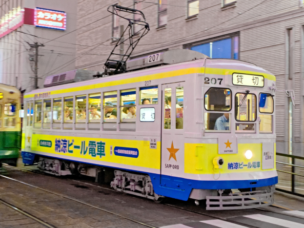 Eine gelbe und blaue Straßenbahn auf einer Stadtstraße in der Nacht mit Menschen drinnen, Gebäuden mit Fenstern, Strommasten mit Drähten und Schildern im Hintergrund.