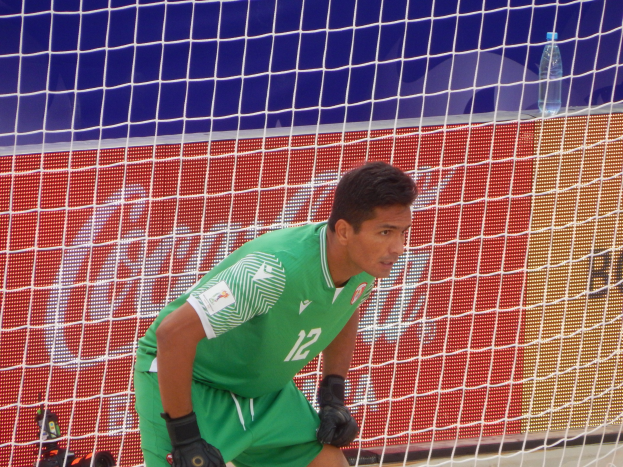 Ein Torwart in grüner Uniform kniet auf dem Boden neben einem Tor mit einem Netz im Hintergrund und einer Flasche rechts daneben.