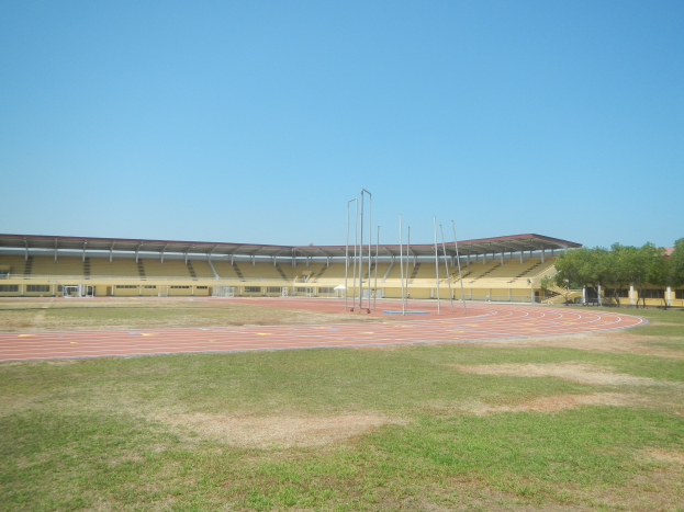 Großes Stadion mit einer zentralen Laufbahn, umgeben von grünem Gras und Bäumen auf der rechten Seite, Pfosten im Hintergrund und einem klaren blauen Himmel.