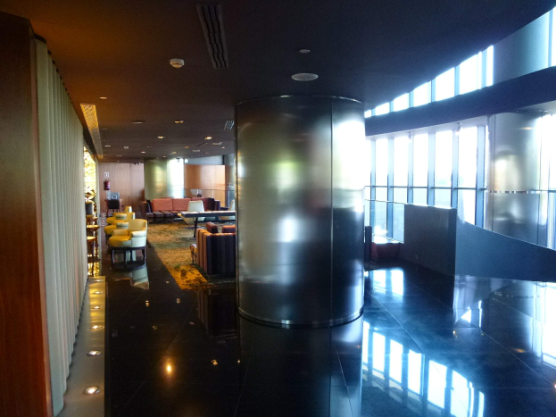 Eine geräumige First-Class-Lounge am Hong Kong International Airport mit großen Fenstern, hohen Decken mit elektrischem Licht, Sitzgruppen einschließlich Stühlen und Sofas, Tischen, einem Teppichboden und einer mittigen Säule.