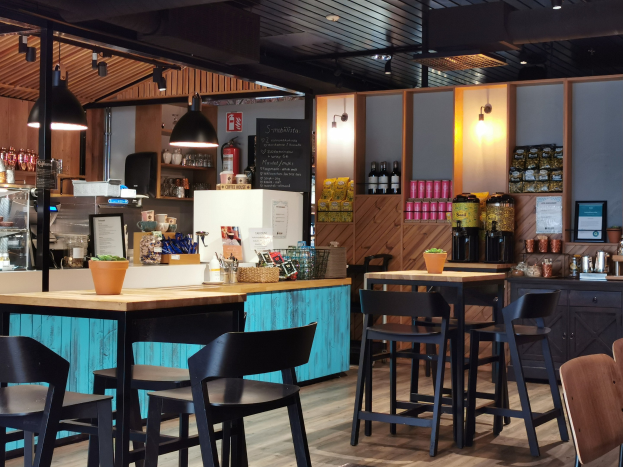 Innenraum eines Restaurants mit Holztischen, Stühlen, einer Topfpflanze auf einem Tisch, einer Schiefertafel, einem Kühlschrank, einem Feuerlöscher, Regalen mit Flaschen, Schränken mit Gegenständen, einem gerahmten Foto an der Wand, Deckenlampen und Lampen.