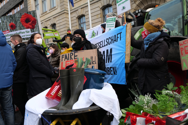 Eine Gruppe von Menschen in Masken mit Protestschildern vor einem Lastwagen, mit einem Tisch voller Gegenstände, Topfpflanzen und einem Gebäude mit Fahne im Hintergrund.