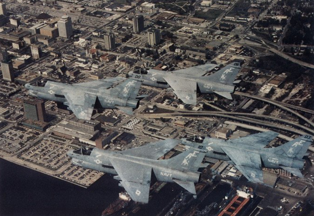 Vier Kampfflugzeuge in Formation über einer Stadt mit sichtbaren Gebäuden, Straßen, Fahrzeugen und Wasser, mit Text am unteren Bildrand.