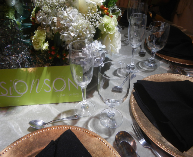 Formaler Esstisch mit goldenen Untersetzern, schwarzen Servietten, Besteck, Gläsern und einem Blumenbouquet, begleitet von einer Texttafel.