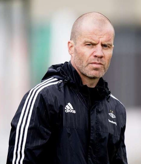 Ein kahlköpfiger Trainer in einer blauen Jacke mit weißen Streifen steht auf einem Fußballfeld, die Hände verschränkt, mit einem ernsten Gesichtsausdruck und einem unscharfen Hintergrund.