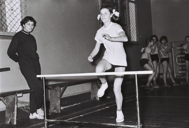 Schwarzes und weißes Foto einer Frau in der Luft, die über ein Hindernis springt, in einer Turnhalle mit Zuschauern und Bänken im Hintergrund.