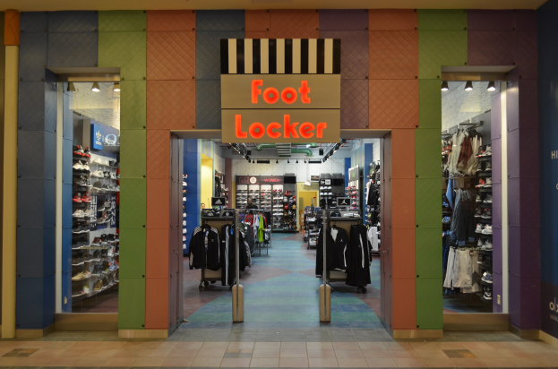 Innenansicht eines Foot Locker-Schuhgeschäfts mit Kleidung an Bügeln und Gestellen unter Deckenbeleuchtung, wobei der Geschäftsboden sichtbar ist.