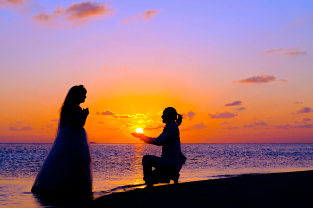 Braut und Bräutigam stehen am Strand bei Sonnenuntergang, Braut in einem weißen Kleid, Bräutigam kniend, während er ein Objekt hält, leuchtender Himmel mit orangefarbenen, pinken und violetten Tönen, tiefblaues Wasser im Hintergrund.