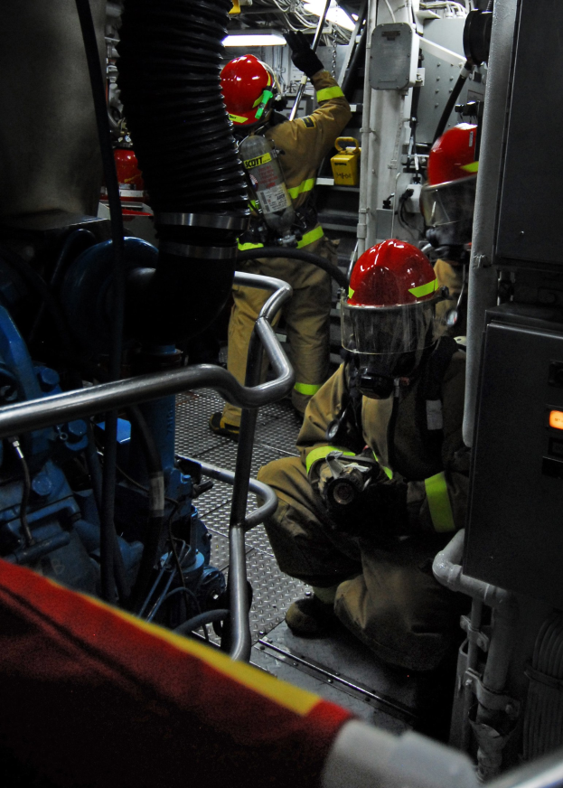 Feuerwehrleute in Helmen und Schutzausrüstung arbeiten an einem Feuerlöschfahrzeug mit Rohren und Gegenständen im Hintergrund und einer Lampe oben.