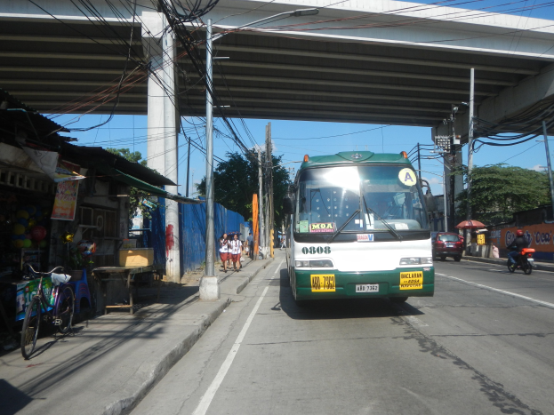 Ein grüner und weißer Bus fährt auf einer Straße mit Strommasten und -leitungen vorbei an Geschäften, Fußgängern und parkenden Fahrrädern in der Nähe einer Brücke, mit Bäumen und einem klaren Himmel im Hintergrund.