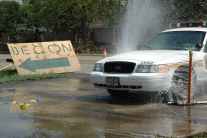 Ein Polizeiwagen fährt durch eine Pfütze, mit einer Tafel, einem Verkehrskegel und anderen Gegenständen am Boden, mit Bäumen, Gebäuden und einem klaren blauen Himmel im Hintergrund.