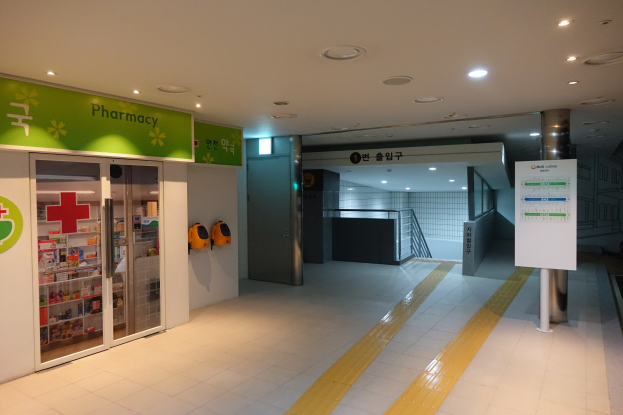 Apothekeneingang mit einem "Apotheke"-Schild auf der linken Seite, einer Tafel mit Text auf der rechten Seite, beleuchteten Lampen darüber und sichtbaren Gegenständen im Hintergrund.