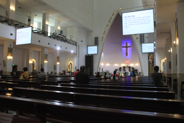 Eine große Gruppe von Menschen auf Kirchenbänken in einer Kirche, mit Bänken, Säulen, Geländern, Bildschirmen, Lampen, Texttafeln, Statuen, Blumenbouquets im Hintergrund und der Decke oben.