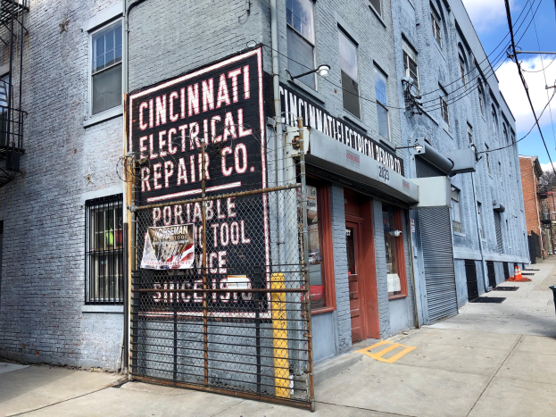 Außenansicht des Cincinnati Electrical Repair Co. Gebäudes mit einer Schautafel, einem Metallzaun, einem Müllcontainer, Strommasten, Kabeln, Bäumen und einem bewölkten Himmel.