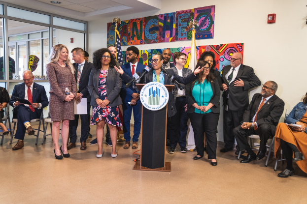 Gruppe von Menschen vor einem Rednerpult mit einem Mikrofon und einer Tafel mit der Aufschrift "Sancho City Councilman Joins City Council", mit sitzenden und stehenden Gästen, Flaggen, Feuerlöscher und Deckenlampen im Hintergrund.