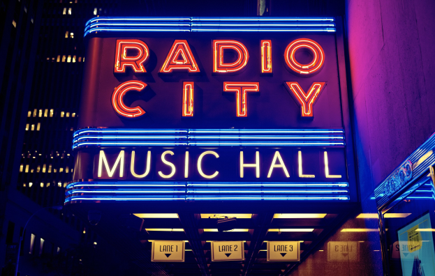 Hell erleuchtetes Radio City Music Hall in New York City mit hohen umliegenden Gebäuden und einem "Radio City"-Schild sowie zusätzlichen Schildern auf der rechten Seite.