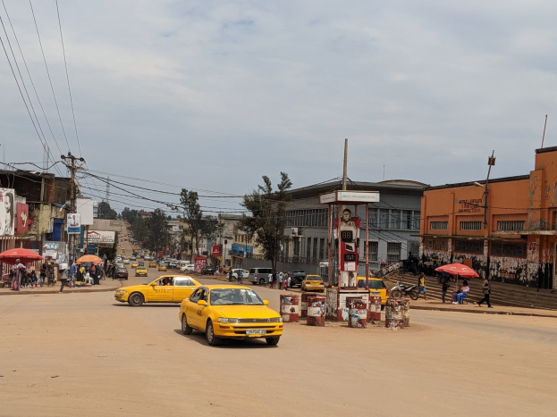 Ein gelbes Taxi fährt eine belebte Stadtstraße entlang, gesäumt von Gebäuden, Fußgängern mit Regenschirmen auf dem Gehweg und Strommasten im Hintergrund bei bewölktem Himmel.