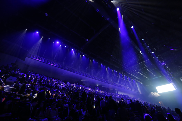 Eine große Menschenmenge in einem Konzertsaal, die einer Bühne mit Deckenleuchten und einem Bildschirm auf der rechten Seite zugewandt ist.