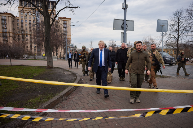 Eine Gruppe von Menschen geht auf einer Straße neben Absperrband, einige halten Waffen, mit Gebäuden, Bäumen, Pfählen, Brettern, Fahrzeugen und einer Barrikade im Hintergrund.