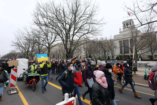 Ein großer Protestmarsch mit Menschen, die eine Straße in Washington, D.C. entlanggehen, einige halten Schilder und fahren Fahrräder, mit Bäumen und einem klaren blauen Himmel im Hintergrund.