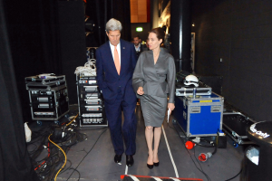 Angelina Jolie und John Kerry auf einem Boden umgeben von einer Gruppe von Menschen, verschiedenen Geräten, Kabeln, einem Helm auf einem Wagen, einem Müllbehälter, einem Vorhang, einer Säule, einem Schild und einer Decke mit Deckenleuchten.