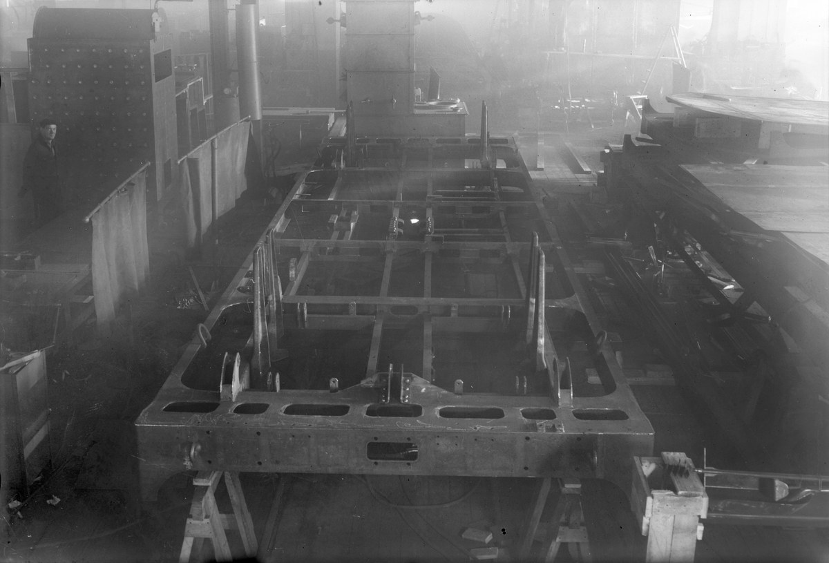 Schwarz-weiß-Foto eines Autos in der Fertigung in einer Fabrik, mit einer Person links und verstreuten Werkzeugen/Materialien.