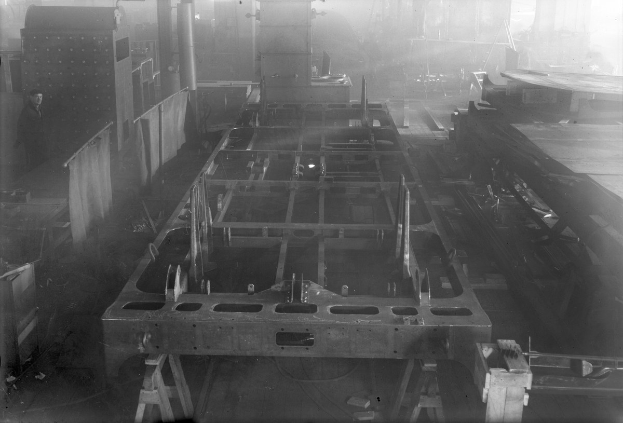 Schwarz-weiß-Foto eines Autos in der Fertigung in einer Fabrik, mit einer Person links und verstreuten Werkzeugen/Materialien.