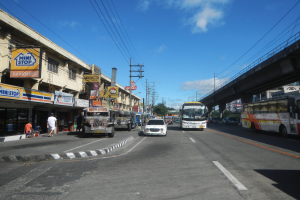Eine belebte Stadtstraße mit Autos, Bussen, Fußgängern, Gebäuden, einer Brücke, Bäumen und einem bewölkten Himmel.