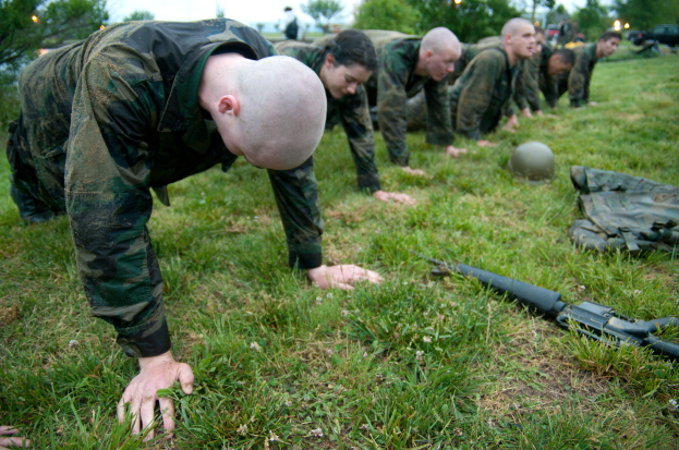 Eine Gruppe von Männern in Militäruniformen, die Liegestütze auf dem Gras machen, mit einer Waffe, einem Helm und anderen Gegenständen drumherum, unter einem klaren blauen Himmel mit Bäumen, Lichtern und Fahrzeugen im Hintergrund.