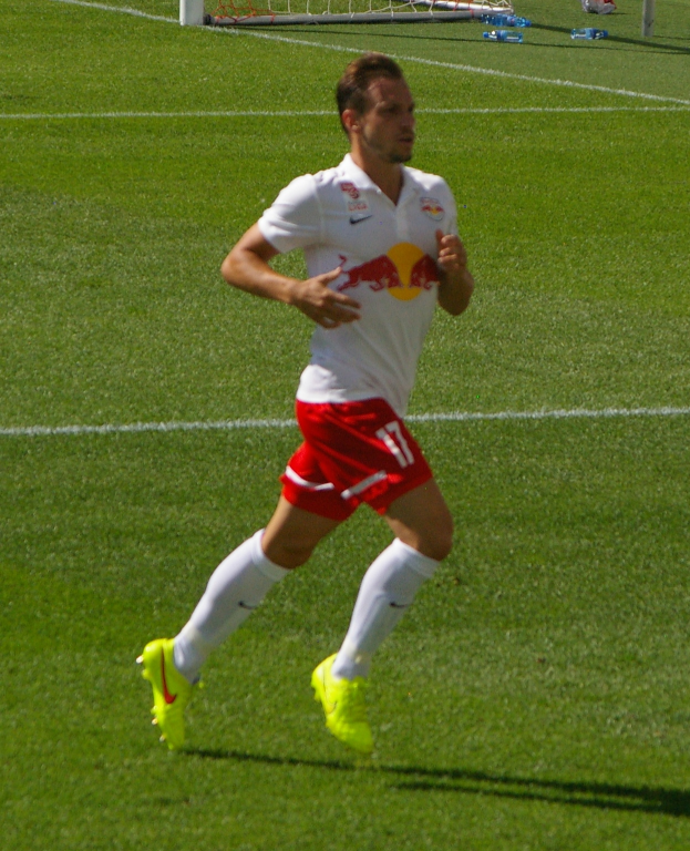Ein Mann in einem weißen Hemd und roten Shorts rennt auf einem Fußballfeld, im Hintergrund ist ein Torpfosten zu sehen und ein paar Menschen stehen in der Nähe, die Fußballschuhe tragen.