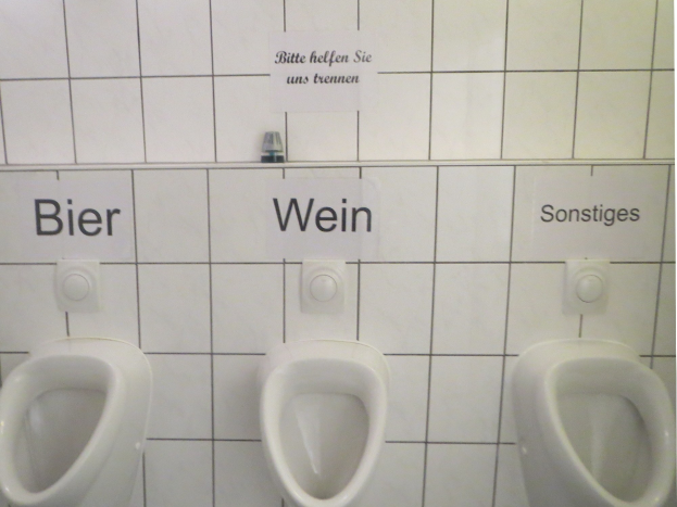 Drei weiße Toiletten an einer Wand montiert mit Schildern darüber, die jeweils "Bier Wein" und "Sonstiges" lesen.