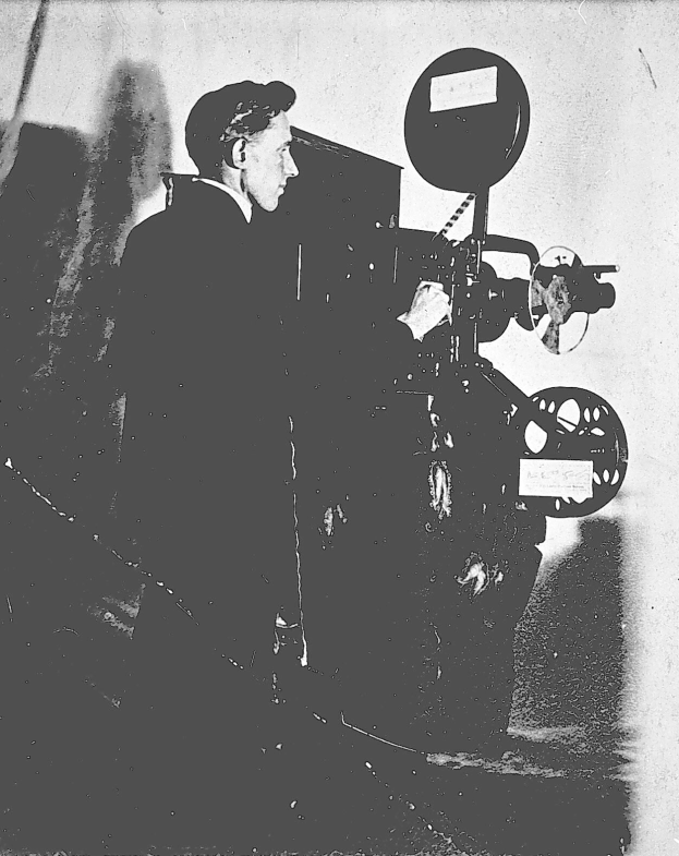 Ein Mann in einem Anzug steht neben einem alten Filmprojektor und sieht konzentriert aus.
