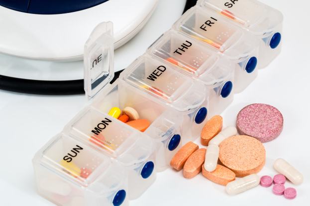 Weißer Tisch mit Pillenschachtel, die Pillen enthält, und einem Stethoskop daneben. Die Pillenschachtel ist als Medikamentenbehälter beschriftet.