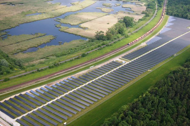 Luftaufnahme einer Solar-Farm mit Panelen, umgeben von Bäumen, Gras, Wasser und einer nahen Bahn.