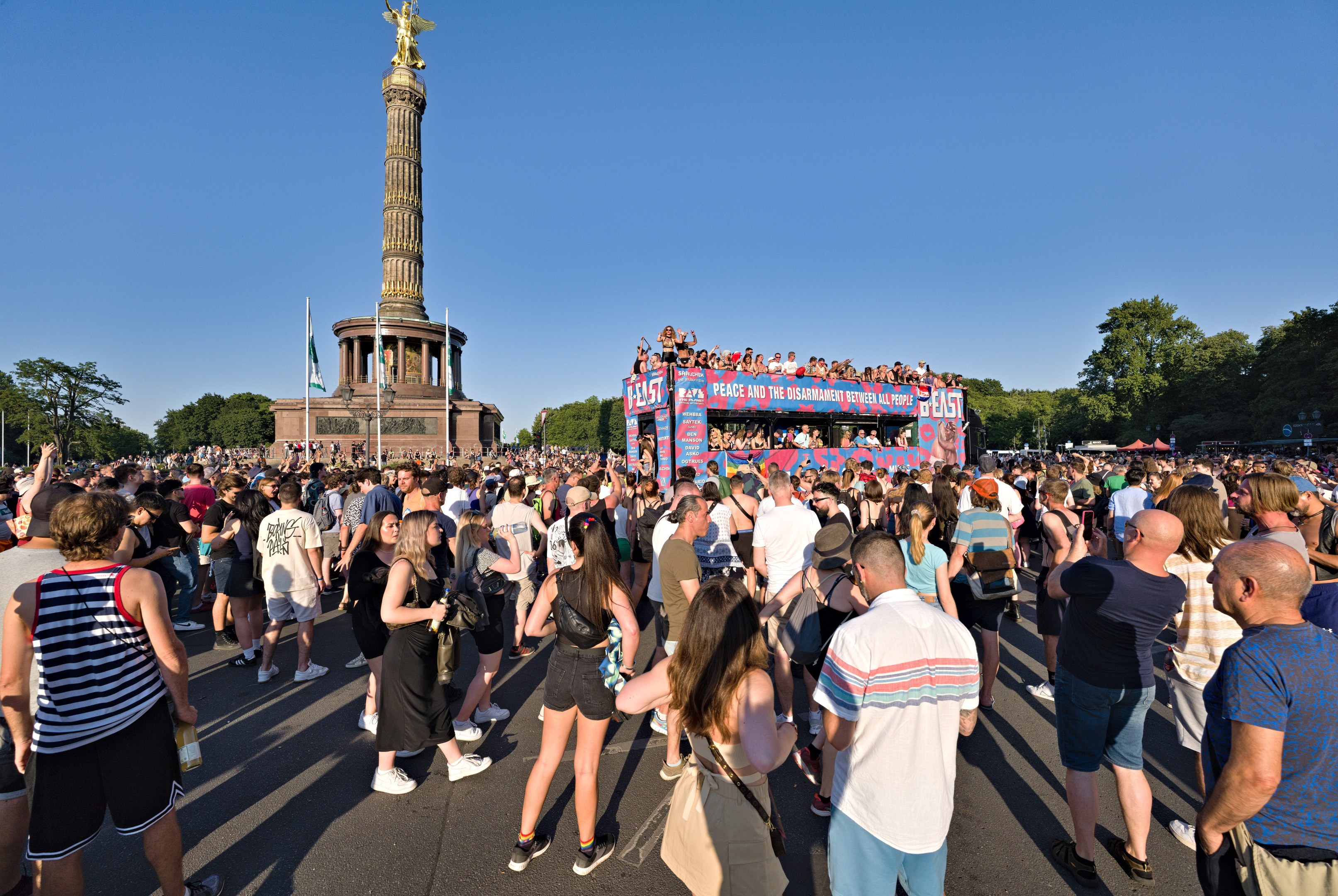 Große Menschenmenge versammelt sich vor einem Denkmal in Berlin mit Fahnen, einem säulenumgebenen Gebäude mit Statue und Inschrift, Bäumen und einem klaren blauen Himmel.