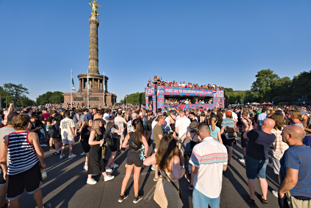 Große Menschenmenge versammelt sich vor einem Denkmal in Berlin mit Fahnen, einem säulenumgebenen Gebäude mit Statue und Inschrift, Bäumen und einem klaren blauen Himmel.