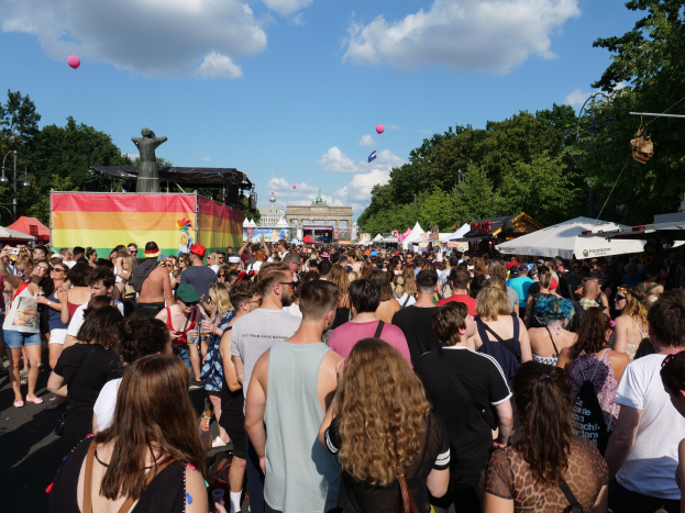 Eine große Menschenmenge geht eine Straße mit Zelten, Bäumen, Pfählen, Lichtern und einer Statue entlang, mit Gebäuden und einem bewölkten Himmel mit Ballons im Hintergrund während des Christopher Street Day in Berlin.