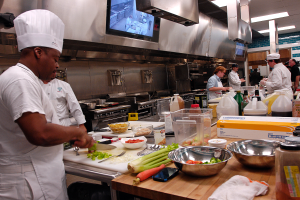 Eine Gruppe von Köchen in einer Küche, die Essen auf einem Schneidbrett zubereiten, mit einem Koch im Vordergrund in einer weißen Schürze und einer Mütze, der ein Messer hält und von Utensilien, Schüsseln, Gemüse, einem Handy, Flaschen und anderen Gegenständen umgeben ist, mit einem Fernseher und Deckenlampen im Hintergrund.