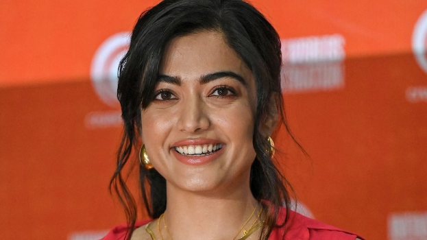 Eine Frau in einem roten Kleid mit einer eleganten Hochsteckfrisur und dezentem Make-up steht strahlend da, trägt schwarze High Heels und hat ein hellgelbes Banner mit der Aufschrift 'Die besten Schönheits Looks von Sonam Kapoor beim Cannes Film Festival' im Hintergrund.