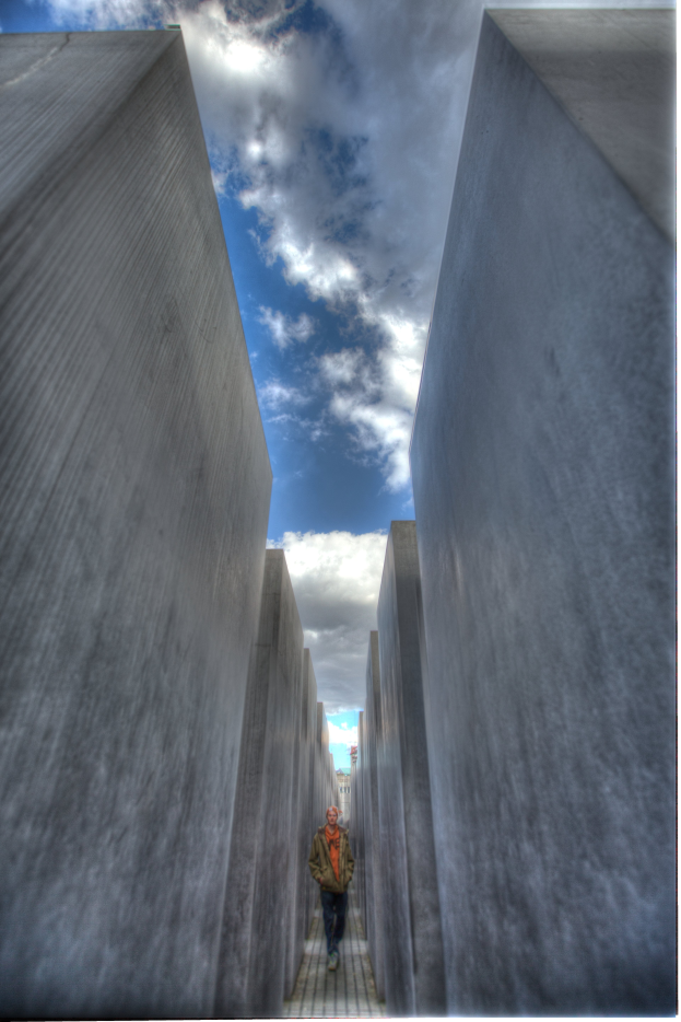 Ein Mann geht durch das Holocaust-Mahnmal in Berlin, Deutschland, mit hohen Betonsteinen auf beiden Seiten und einem bew"{o}lktem Himmel im Hintergrund.