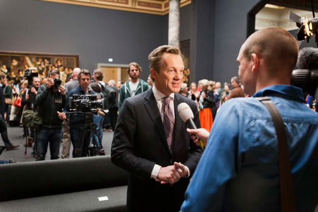 Ein Mann im Anzug und mit Krawatte spricht mit einem Reporter, der ein Mikrofon hält, vor einer Menge von Menschen, einige halten Kameras, im Hintergrund eine Couch und eine Wand mit einem Foto, Lampen und einer Säule.