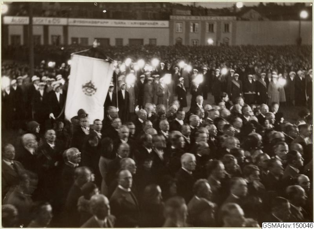 Schwarzes und weißes Foto einer großen Menge vor einem Gebäude, mit einer Person, die eine Fahne hält, in der Mitte, Pfosten und Lichter im Hintergrund und Text, der unten "Gustav Klimts Rede an der Berliner Mauer" lautet.