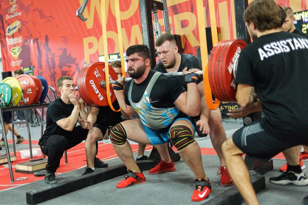Eine Gruppe von Männern in einem Fitnessstudio, die Kniebeugen mit Langhanteln machen, umgeben von verschiedenen Fitnessgeräten und Bannern mit der Aufschrift "Powerlifting-Weltmeisterschaften 2019".