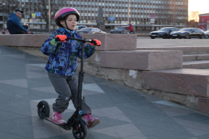 Ein junger Junge fährt mit einem Helm und Handschuhen auf einem Gehweg auf einem Scooter, mit verschiedenen städtischen Elementen und einem klaren blauen Himmel im Hintergrund.