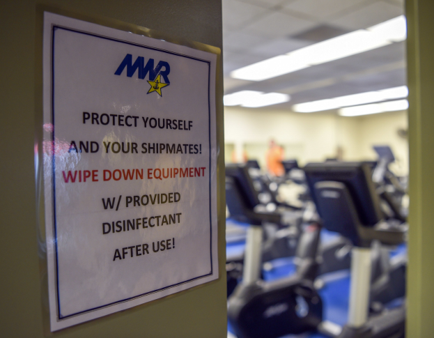 Ein Türschild mit der Aufschrift "Schützen Sie sich und Ihre Schiffskameraden" mit Laufbändern und Deckenbeleuchtung im Hintergrund, das Bild leicht unscharf, um Bewegung anzudeuten.