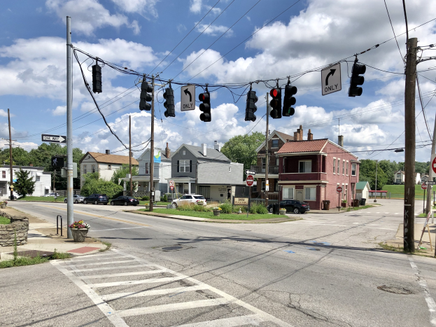 Stadtstraße mit Zebrastreifen, Ampeln, Fahrzeugen, Strommasten, Schildern, blühenden Pflanzen, Gras, Bäumen, Gebäuden und bewölktem Himmel.